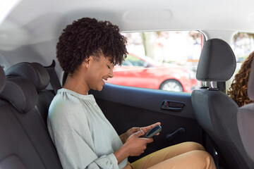 Smiling black woman using smartphone while sitting in the back seat of a car. Young woman checks...