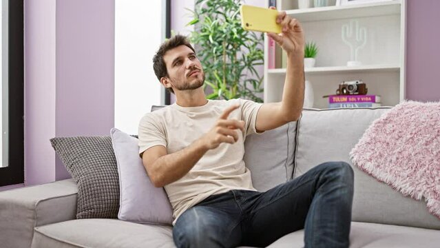 Hispanic man taking selfie with smartphone on couch in cozy living room interior