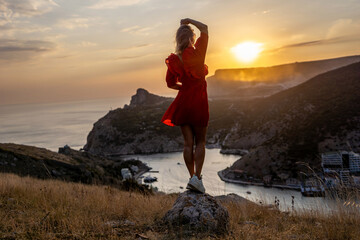 Happy woman standing with her back on the sunset in nature in summer with open hands posing with mountains on sunset, silhouette. Woman in the mountains red dress, eco friendly, summer landscape 