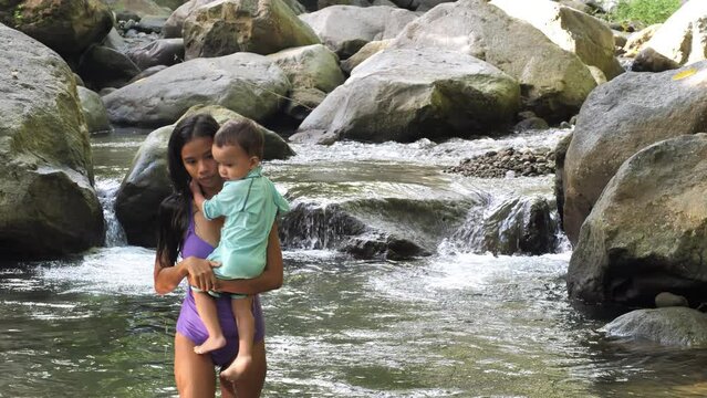 Woman with little baby gets out of river against large rocks