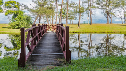 Obraz premium A pedestrian wooden bridge with railings spans the river. Green grass and trees grow on the lawn. The ocean is ahead. Clouds in the blue sky. A mirror image in calm water. Malaysia. Borneo. 