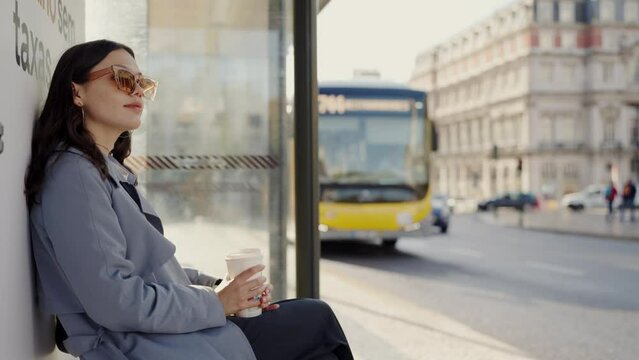 Young female in relaxed mood sitting on bench of bus stop while waiting transport. Stylish woman in trendy sunglasses on bus shelter outdoors. City lifestyle
