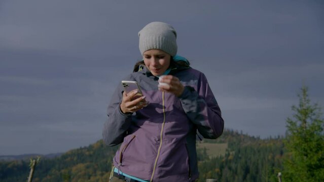 Female Caucasian tourist takes selfie in front of beautiful scenery. Adult woman uses her phone to watch photos. Hiker during expedition in mountains in autumn. Tourism and active leisure concept.
