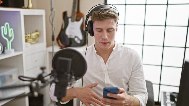 Confident, young caucasian man is a multitalented radio reporter, dishing out hot news through the microphone, reading from his smartphone screen on the broadcast stage of an indoor radio studio