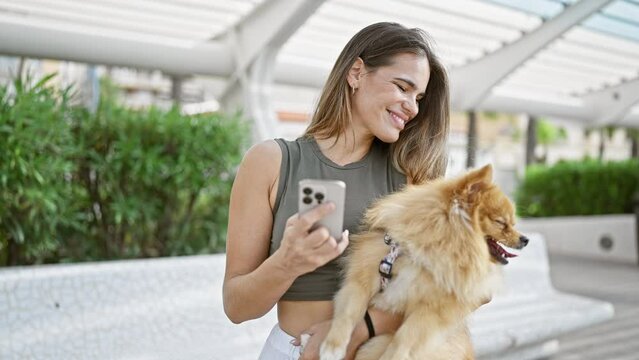 Young, confident hispanic woman joyfully smiling, hugging her beautiful pet dog, while cheerfully texting on her smartphone at the park.