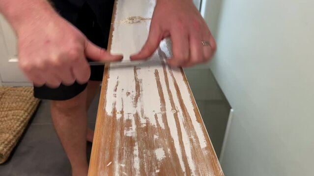 Close up of a man's hands scraping white paint off the top of a wooden bannister with a steel ruler