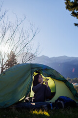 child sits in a tent against the backdrop of the mountains and drinks water from a bottle. boy in a sleeping bag. camping and trekking with children.