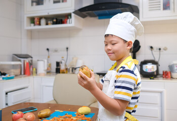 Cute happy smiling Asian 8 years old boy child wearing chef hat and apron having fun preparing,...