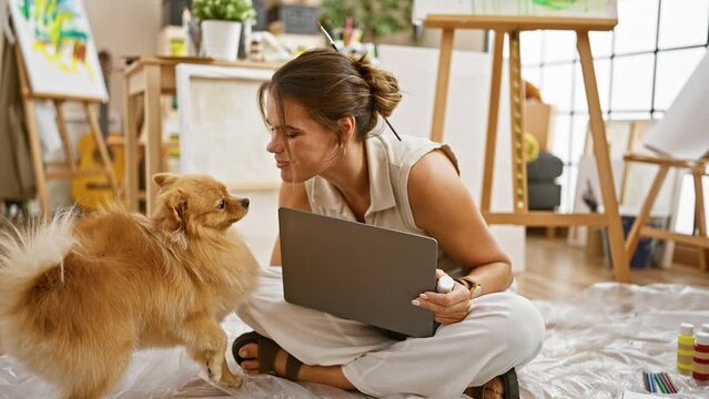 Smiling young hispanic woman artist kisses her beloved pet dog, sitting on studio floor, engaging in her artistry through a laptop, creating joy in the learning process.