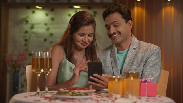 Happy Indian couples at candlelight dinner using mobile phone at restaurant - concept of social media sharing, online app or application and anniversary celebration.