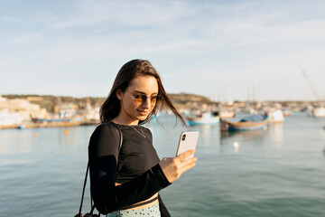 Happy smiling romantic woman with flying hair wearing glasses and black top is using smartphone against ocean with phone