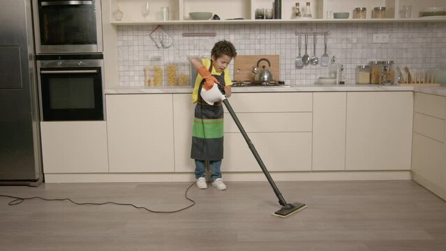 Cute school age African American boy in apron doing household chores, washing and cleaning kitchen floor using steam cleaner while helping with housework at home.