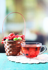 Forest berries in wicker basket and glass cup with red fruit tea, on wooden table, on bright background - obrazy, fototapety, plakaty