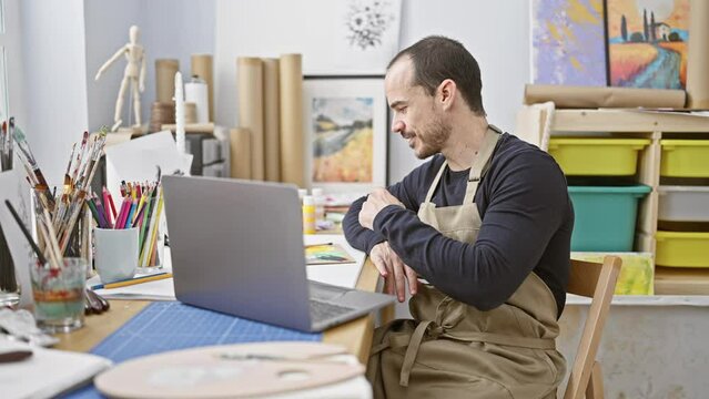 A bearded man in a bib apron smiles during a video call in a creative art studio, surrounded by paintings and art supplies.