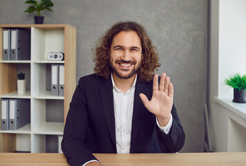 Cheerful businessman says hello at the start of an online business meeting. Video portrait of a happy long haired young man in a suit waving his hand, greeting his colleagues and smiling