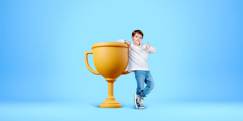 Boy with thumb up standing near gold cup on empty blue background