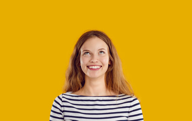 Fototapeta premium Thoughtful dreaming pensive reflective smiling young woman with wavy long hair in striped sweatshirt looking up on yellow background. Sincere emotions concept. Banner for advertisement, marketing.