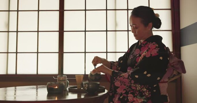 Japanese woman, pot and tea cup for ceremony with health, wellness or liquid to relax at tearoom table. Lady, person and pouring with drink for culture, detox or peace with indigenous clothes in home