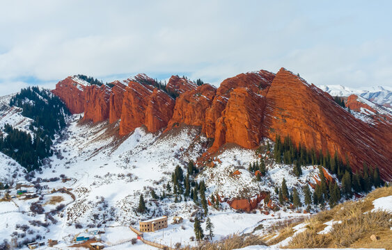 Red cliffs known as Jeti-Oguz's Seven Bulls, with snow and greenery, under a semi-cloudy sky.