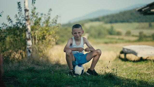 Blond Countryside Boy Smiling While Sitting on a Ball and Tearing the Grass. Concept of Future Athlete, Aspiring Footballer, Summer Olympics, UEFA EURO, FIFA World Cup