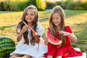 Little Caucasian cute girls eating watermelon while picnic at park. Children sisters sitting on green grass together in summer. Family time