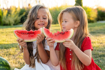 Little Caucasian cute girls eating watermelon while picnic at park. Children sisters sitting on green grass together in summer. Family time