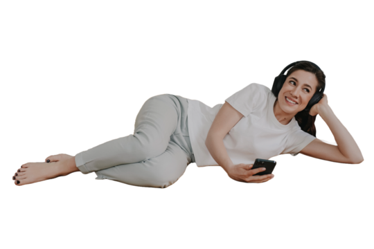 Indoor shoot of cheerful young Spanish woman laying on sofa holding smartphone looking aside against transparent background, listening music using headphones, leans on hand, toothy smiling