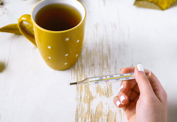 Top view of female hands holding cup of tea and thermometer on shabby table with yellow scattered leaves . autumn composition