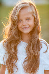 Portrait of smiling little girl walks in the rays of a sunset in a flowering meadow, enjoying the summer, warmth, flowers, freedom. Child in white dress is having fun outdoors.