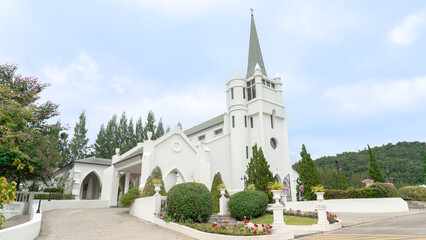 Fototapeta na wymiar Location photos Tall European Church The pitched roof has a cross that sends devotion to God in heaven. The building is white and clean, like a land of paradise. Colorful flower bushes decorate the fr