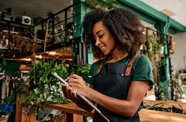 Running of own business. Side view of african woman wearing green apron writing on clipboard while doing inventory in floral boutique. Female shop owner with dark curly hair inspecting supplies.