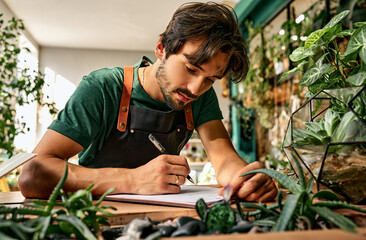 Inventory at floristic shop. Focused caucasian man leaning on desk and writing on clipboard while working at modern store. Bearded florist running successful nursery with green domestic plant. - obrazy, fototapety, plakaty