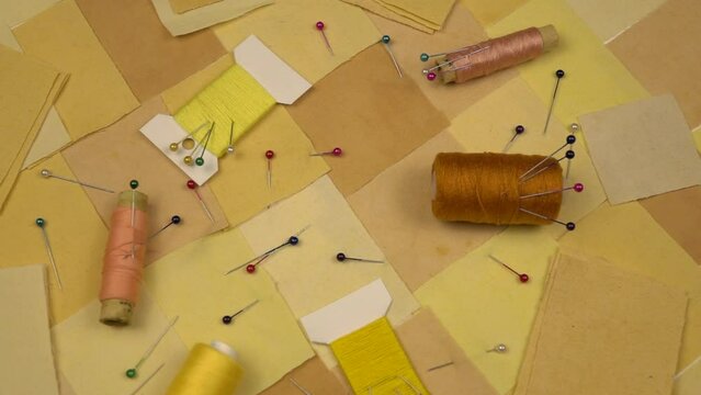 Accessories for patchwork sewing. Needles, tailor's pins, spools of thread, fabric blanks lie on a textile in patchwork style in yellow tones, slow motion