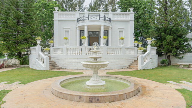 Location photos A three-tiered, circular white fountain with a stone tile floor. In the middle of the green grass The back is a luxurious European-style building decorated with vases, large trees, 
