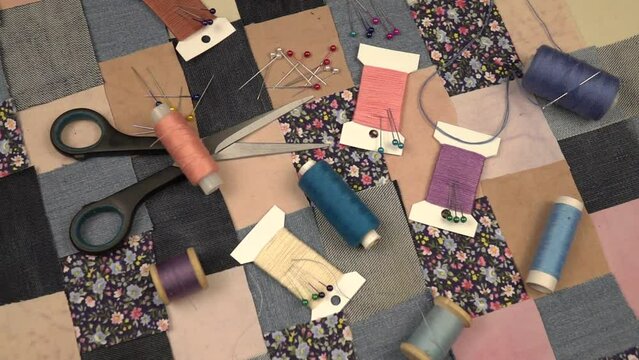 Accessories for patchwork sewing. Needles, tailor's pins, spools of thread, scissors lie on a textile in patchwork style with printed cotton fabric and denim elements in blue tones, slow motion