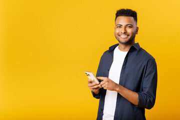 Online communication. Handsome happy indian man using smartphone standing isolated on yellow, arab...