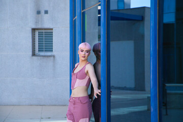 Young gay guy with pink hair and make-up leans against a glass and mirrored office building. The boy is dressed in pink. Concept of equality and LGBTQ rights.