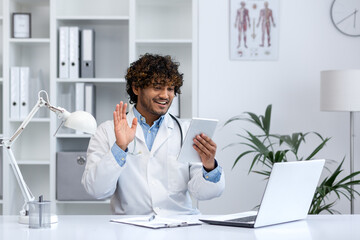 Young successful doctor using tablet computer for video call, man in white medical coat clinic worker consulting patients remotely sitting at desk inside office.