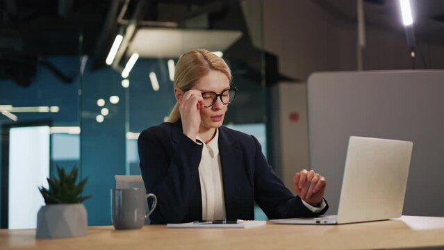 Overtired businesswoman in a blue suit and glasses in the office. Woman feels tired from work, takes off glasses rubs temples to ease headache. Female person suffers from headaches and lack of sleep.