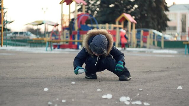  Child toddler caucasian ukrainian boy in warm winter clothes playing with a piece of ice on a cold playground