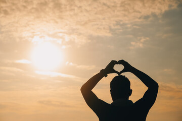 .Hand heart frame shape silhouette made against the sun & sky of a sunrise or sunset on a mountain.