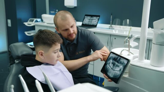 Pleasant friendly dental expert showing x-ray image of teeth on gadget to young patient during dental checkup. Boy in dentist chair, fully equipped office. High quality 4k footage
