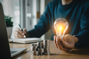 Businessman holding and putting lightbulb on coins stack on table for saving energy and saving money concept. - obrazy, fototapety, plakaty