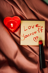 A note with text love yourself drawing with red lipstick on a silk terracotta silk. A burning...