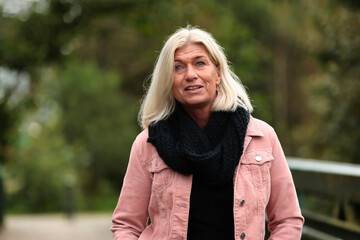 Obraz premium Woman blonde in her fifties with a pink denim jacket, blue jeans with a ripped black sweater and a black scarf, portrait on a bridge.