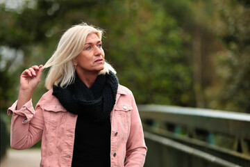 Woman blonde in her fifties with a pink denim jacket, blue jeans with a ripped black sweater and a black scarf, portrait on a bridge.