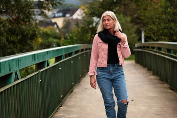 Woman blonde in her fifties with a pink denim jacket, blue jeans with a ripped black sweater and a black scarf, portrait on a bridge.