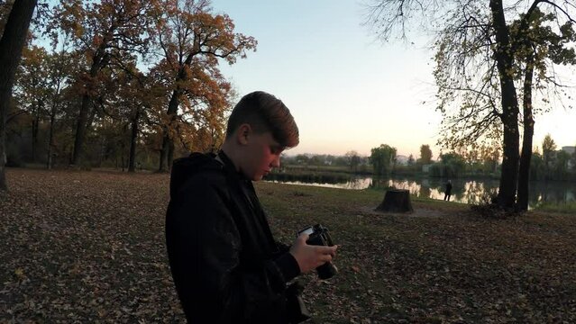 Young caucasian guy boy teen photographer takes pictures in the sunny autumn park among the trees 