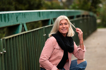 Woman blonde in her fifties with a pink denim jacket, blue jeans with a ripped black sweater and a black scarf, portrait on a bridge.