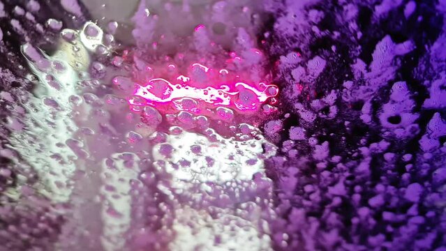 View through the windshield in a car wash with foam and brushes and colored light.